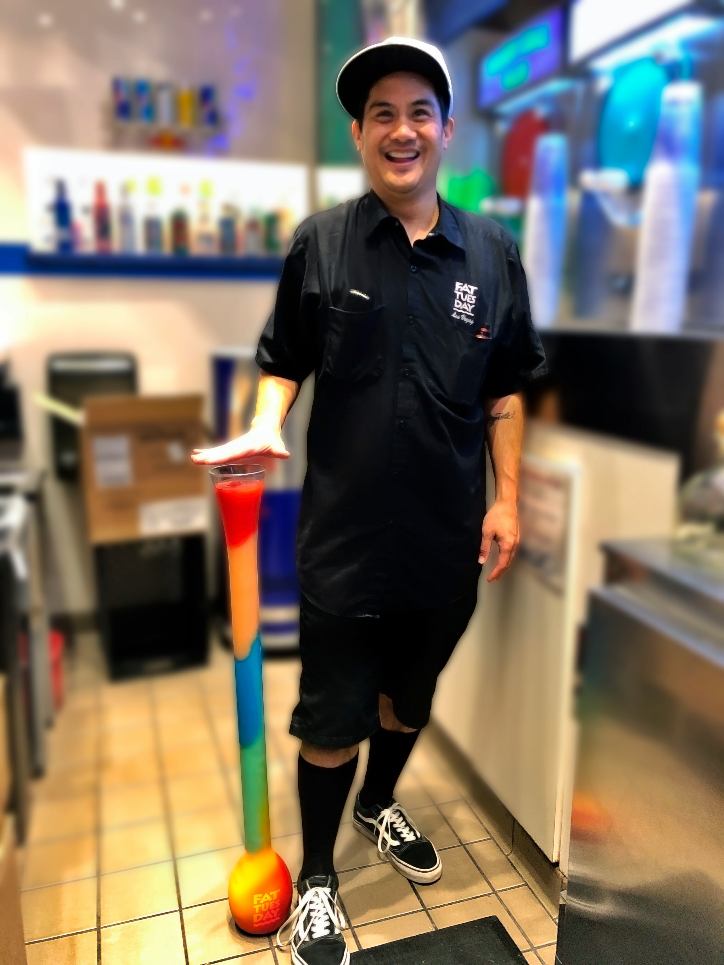 Fat Tuesday bartender looking happy showing off a cocktail with loads of different colours. The cocktail cup is sitting on the floor and goes up to their waist
